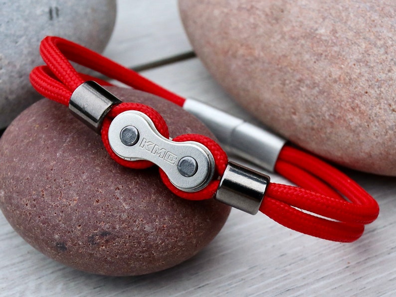 May include: Red rope bracelet featuring a silver-colored bicycle chain link with the letters "KMC". The bracelet has silver-colored metal end caps and a magnetic clasp. The bracelet is displayed on a wooden surface with rocks in the background.