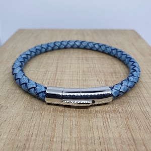 May include: A braided blue leather bracelet with a silver-colored magnetic clasp. The bracelet is circular and sits on a light brown wooden surface. The leather has a textured appearance.