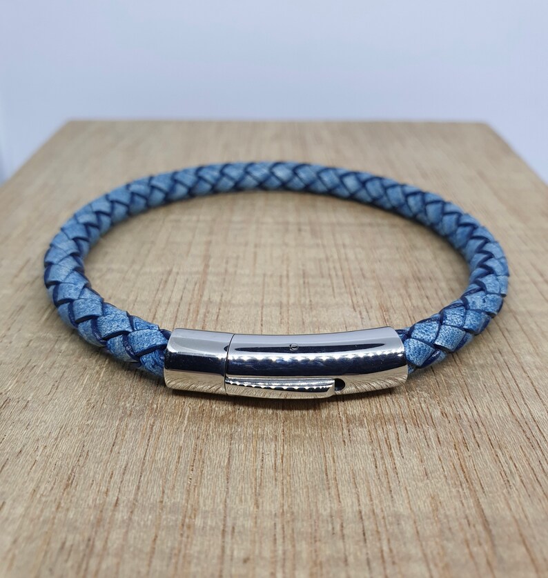 May include: A braided blue leather bracelet with a silver-colored magnetic clasp. The bracelet is circular and sits on a light brown wooden surface. The leather has a textured appearance.
