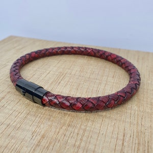 May include: Braided leather bracelet in a deep burgundy color. The bracelet features a woven design and a black magnetic clasp. The bracelet is shown on a wooden surface.