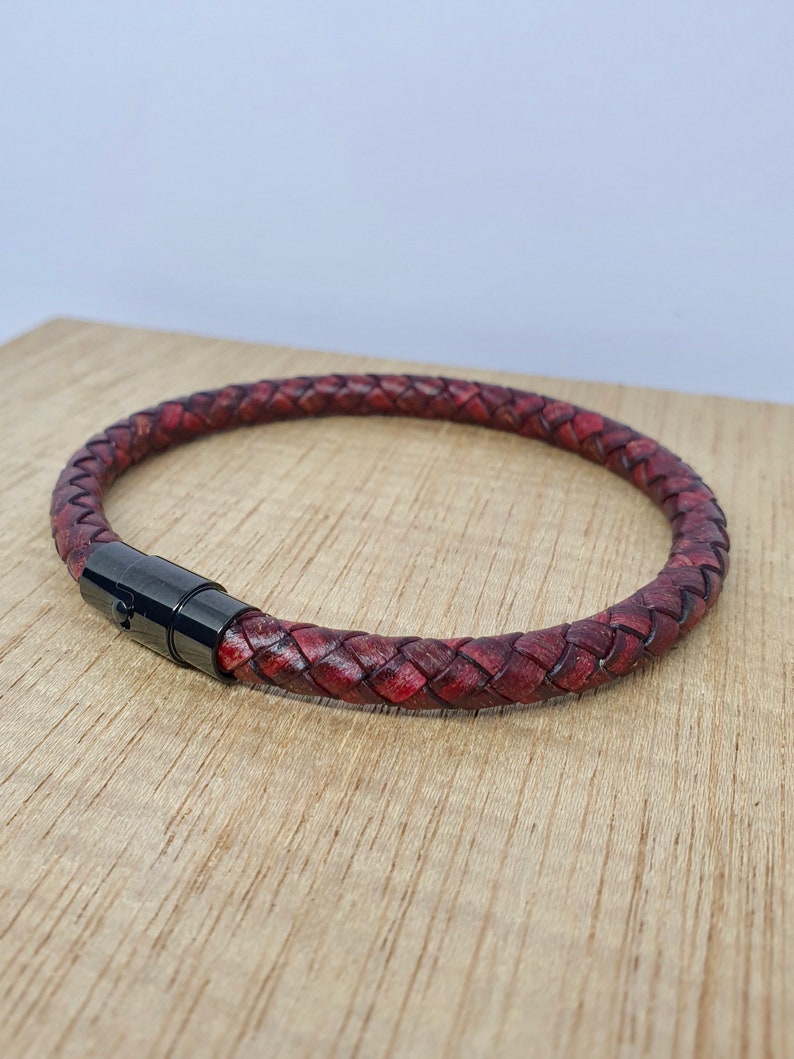 May include: Braided leather bracelet in a deep burgundy color. The bracelet features a woven design and a black magnetic clasp. The bracelet is shown on a wooden surface.