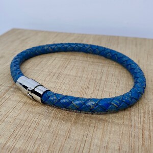 May include: A blue braided leather bracelet with a silver magnetic clasp. The bracelet is circular and appears to be made of a distressed leather material. The bracelet is resting on a light brown wooden surface.