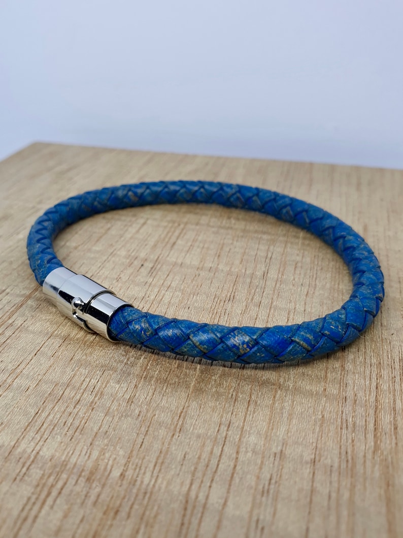 May include: A blue braided leather bracelet with a silver magnetic clasp. The bracelet is circular and appears to be made of a distressed leather material. The bracelet is resting on a light brown wooden surface.