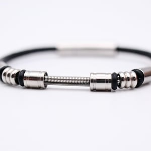 May include: A black leather bracelet with silver metal detailing. The bracelet has a central spring design and cylindrical clasps. The design includes black rubber rings and a silver bar. The bracelet is set against a white backdrop.