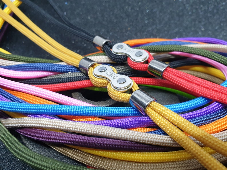 May include: Close-up of a collection of colorful lanyards. Each lanyard features a braided cord in various colors, including yellow, red, blue, and green. The lanyards are connected with a metal clasp and metal ends. The clasp has the letters "KMC" on it.