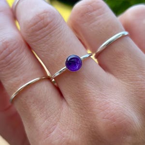 May include: A hand wearing three rings. The central ring features a small, round, deep purple gemstone set in a silver band. Two thinner, plain silver bands flank the gemstone ring. The background is blurred green.