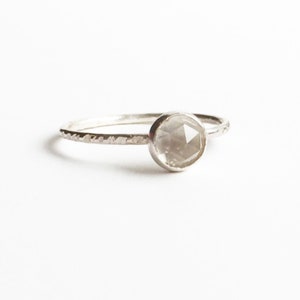 Op de afbeelding: Een zilveren ring met een ronde, facetgeslepen, lichtgrijze edelsteen. De band heeft een textuurontwerp. De ring staat tegen een witte achtergrond.