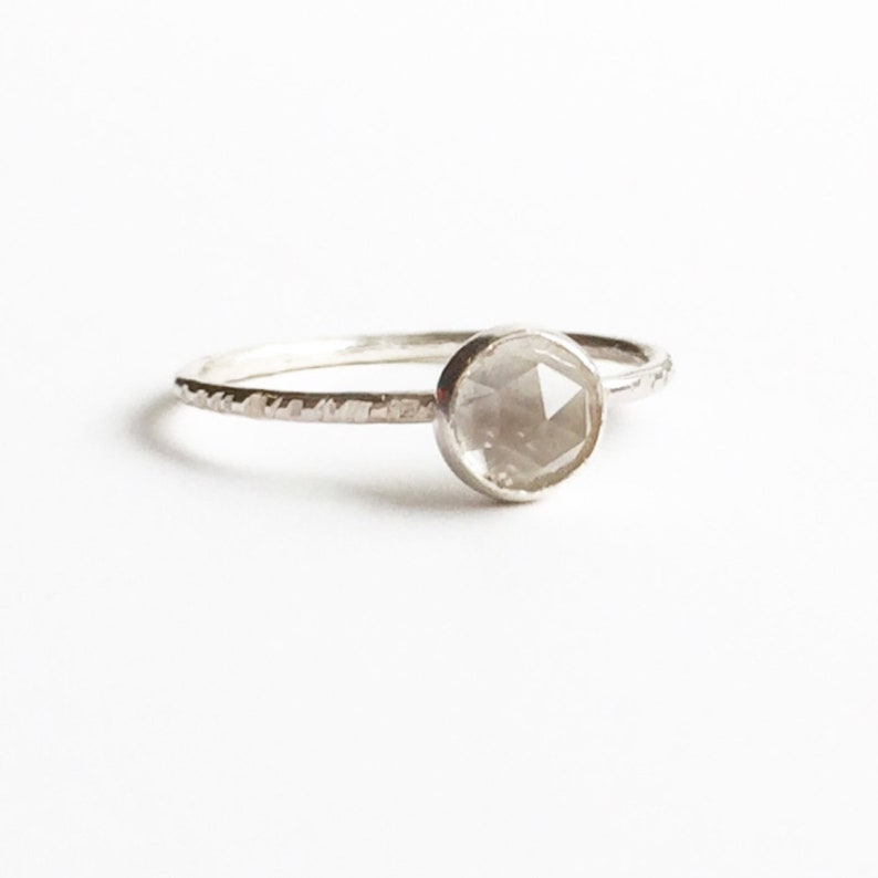 Op de afbeelding: Een zilveren ring met een ronde, facetgeslepen, lichtgrijze edelsteen. De band heeft een textuurontwerp. De ring staat tegen een witte achtergrond.