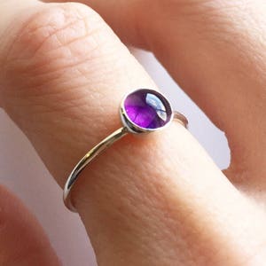 May include: A silver ring featuring a round, vibrant purple gemstone. The gemstone is set in a bezel setting, and the ring has a thin, delicate band. The ring is shown on a finger.