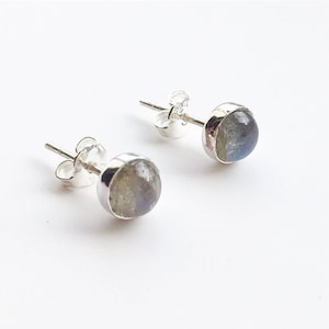 Puede incluir: Un par de pendientes de botón con piedras preciosas redondas e iridiscentes engastadas en metal plateado. Las piedras preciosas muestran una mezcla de tonos grises, azules y blancos. Los pendientes se exhiben sobre un fondo blanco.