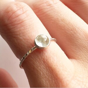 Op de afbeelding: Een zilveren ring met een ronde, facetgeslepen, lichtgroene edelsteen. De band heeft een getextureerd, gehamerd ontwerp. De ring wordt op een vinger getoond.