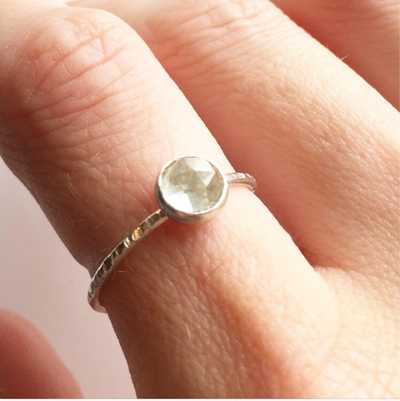 Op de afbeelding: Een zilveren ring met een ronde, facetgeslepen, lichtgroene edelsteen. De band heeft een getextureerd, gehamerd ontwerp. De ring wordt op een vinger getoond.