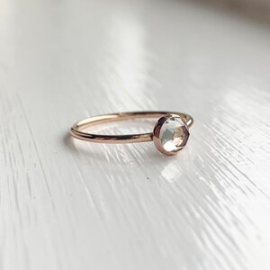 Op de afbeelding: Een delicate gouden ring met een facetgeslepen, heldere edelsteen. De ring heeft een dunne band en een eenvoudig, elegant ontwerp. De ronde edelsteen is gezet in een bezel, wat een minimalistische esthetiek cre&euml;ert.