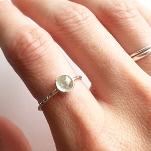 Op de afbeelding: Een delicate zilveren ring met een ronde, lichtgroene edelsteen. De band heeft een textuurontwerp. De ring wordt op een vinger getoond, met een andere zilveren ring op de achtergrond.