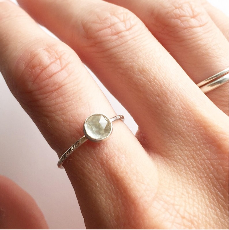 Op de afbeelding: Een delicate zilveren ring met een ronde, lichtgroene edelsteen. De band heeft een textuurontwerp. De ring wordt op een vinger getoond, met een andere zilveren ring op de achtergrond.