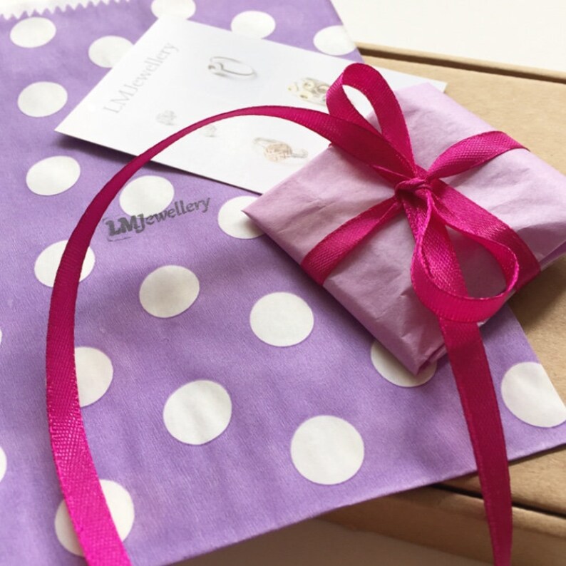 May include: A wrapped gift tied with a bright pink ribbon, resting on a lavender polka dot bag. The gift is wrapped in light pink paper. A card with the text "LMJewellery" is visible.