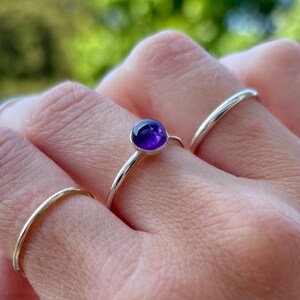 May include: A hand wearing three silver rings. The central ring features a small, round, deep purple amethyst gemstone. The other two rings are simple, thin bands. The background is a blurred green.