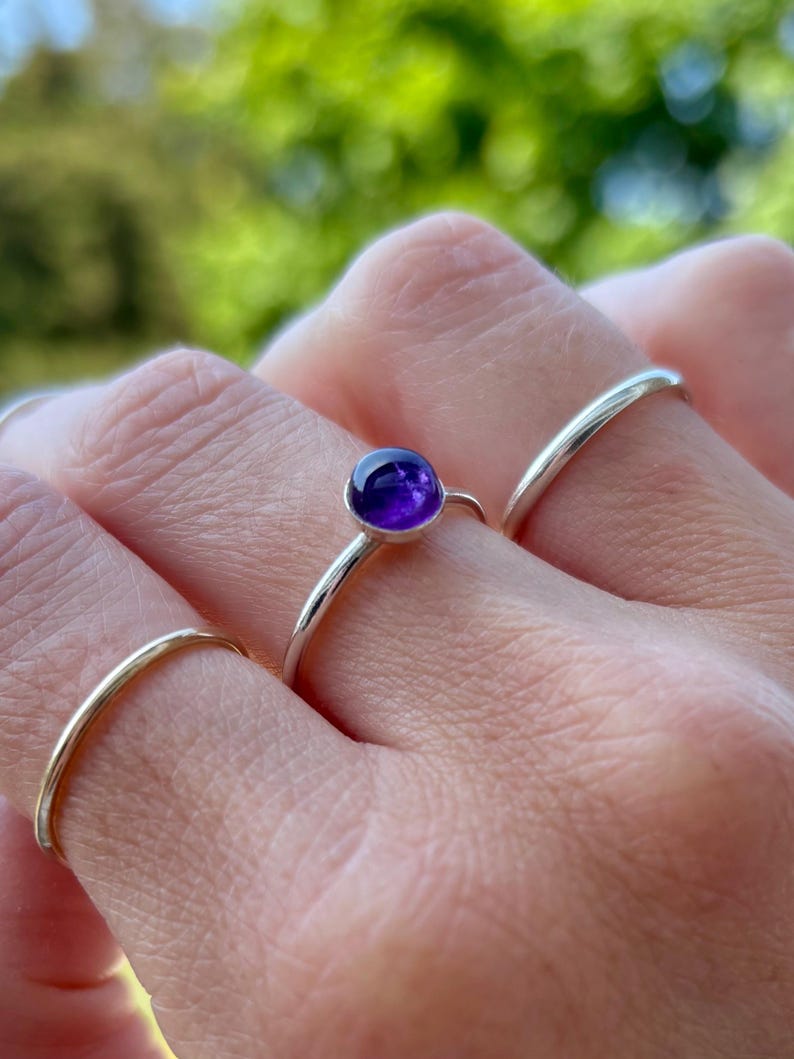 May include: A hand wearing three silver rings. The central ring features a small, round, deep purple amethyst gemstone. The other two rings are simple, thin bands. The background is a blurred green.