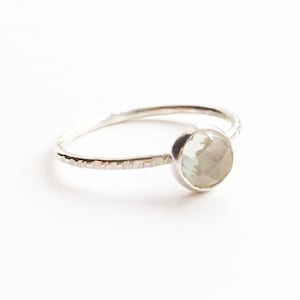 Op de afbeelding: Een zilveren ring met een getextureerde band en een ronde, lichtgekleurde edelsteen. De edelsteen is gezet in een bezel setting en de ring wordt getoond tegen een witte achtergrond. De ring is een sieraad.