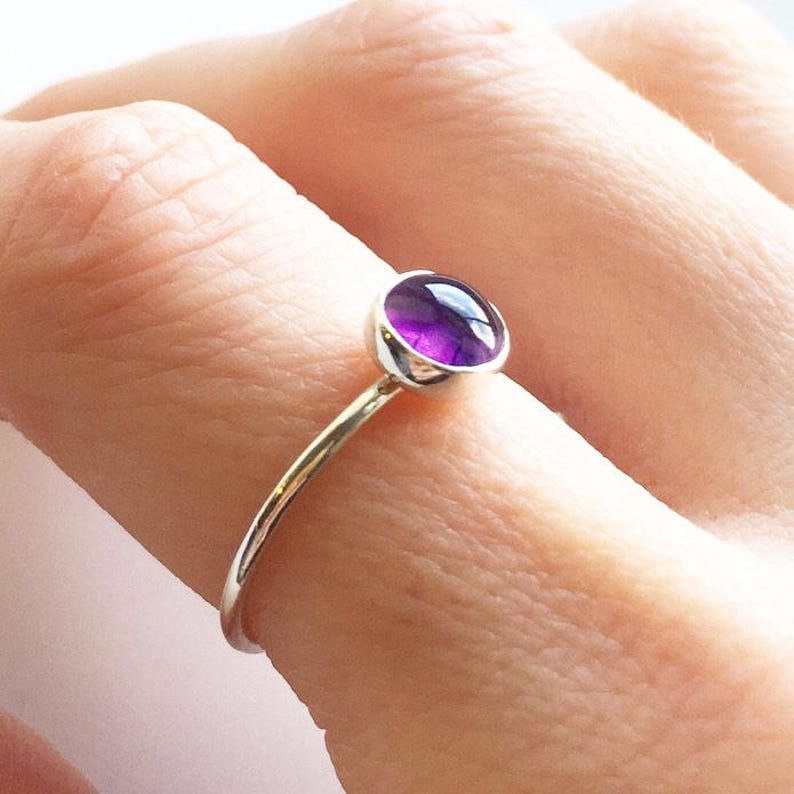 May include: A silver ring featuring a round, vibrant purple gemstone. The gemstone is set in a bezel setting, and the ring has a thin, delicate band. The ring is shown on a finger.