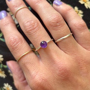 May include: A hand wearing three rings: a gold band, a silver band, and a silver ring with a round, purple gemstone. The background is a black floral print fabric. The nails are painted a light purple color.
