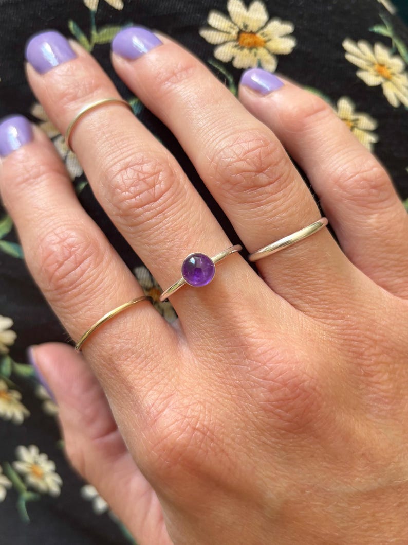 May include: A hand wearing three rings: a gold band, a silver band, and a silver ring with a round, purple gemstone. The background is a black floral print fabric. The nails are painted a light purple color.