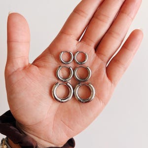 May include: Six silver hoop earrings of varying sizes are displayed in a hand. The earrings are round and have a polished finish. The hand is holding the earrings against a white background. A brown scrunchie and bracelets are visible on the wrist.