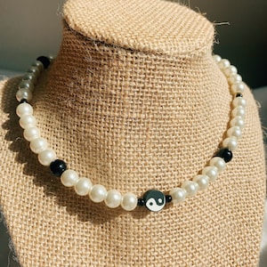 May include: A pearl necklace with a yin-yang pendant. The necklace features white faux pearls and black beads. The yin-yang pendant is black and white. The necklace is displayed on a tan jewelry bust.