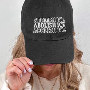 May include: A black baseball cap with white embroidered text that reads "ABOLISH ICE" repeated three times.