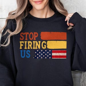 May include: Black sweatshirt with the text "STOP FIRING US" in orange, yellow, and blue. The design includes a stylized American flag. The sweatshirt is a casual garment.