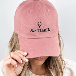 May include: A dusty rose baseball cap with the text "Par-TIMER" and a golf ball and tee graphic. The cap is made of a soft material and has a curved brim. The hat is a golf-themed accessory.