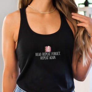 May include: A black racerback tank top featuring a graphic of a small stack of colorful books above the white text that reads: READ. REPEAT. FORGET. REPEAT AGAIN. The tank top is paired with blue denim jeans that have a button and zipper closure.