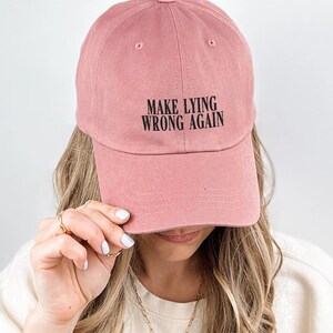 May include: A dusty rose baseball cap with the text "MAKE LYING WRONG AGAIN" in black. The cap is made of a soft material and has a curved brim. The hat is a casual accessory.