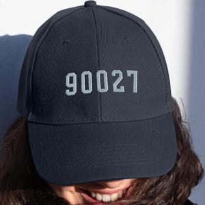 May include: A navy blue baseball cap with the number "90027" embroidered in light gray on the front. The cap is made of a soft fabric and has a curved brim. The person wearing the cap is smiling.