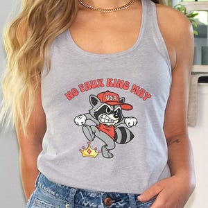 May include: A light gray tank top with a cartoon raccoon graphic. The raccoon wears a red hat with "USA" and kicks a golden crown. The text "NO FAUX KING WAY" is arched above the raccoon.