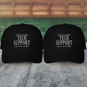 May include: Two black baseball caps with the text "TECH SUPPORT" in white, above a row of stars. The caps are displayed on a light gray wooden surface, with a blurred wooden background.