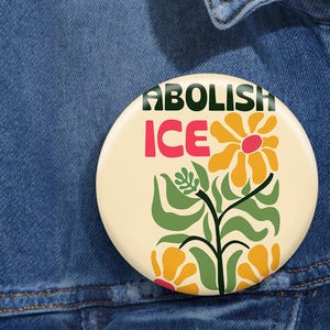 May include: A white button with the text "ABOLISH ICE" in green and pink letters. The button features a stylized floral design with yellow and green flowers.