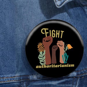 May include: Black and white button with the text "FIGHT authoritarianism" in yellow. The button features three raised fists with brown and peach skin tones and a stylized flower.