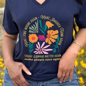 May include: Navy blue V-neck t-shirt with a floral design and text that reads "Make Lying Wrong Again - Make America Think Again - Make Humans Matter Again - Make People Care Again". The shirt is worn by a person.