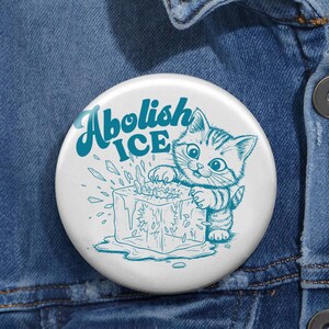 May include: A round pinback button featuring a white background with a teal graphic of a kitten clawing at a melting ice cube. The text "Abolish ICE" is printed in a bold, teal, retro-style font above the kitten. The button is attached to blue denim.