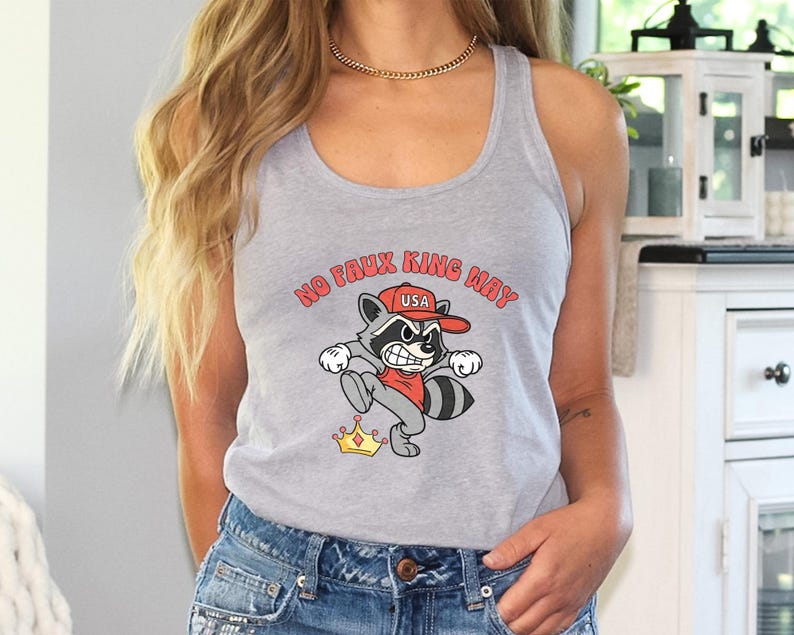 May include: A light gray tank top with a cartoon raccoon graphic. The raccoon wears a red hat with "USA" and kicks a golden crown. The text "NO FAUX KING WAY" is arched above the raccoon.