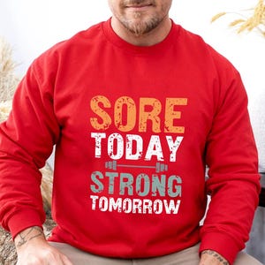 May include: Red long-sleeve sweatshirt with the text "SORE TODAY STRONG TOMORROW" in a vintage style font. The text is layered with a graphic of a barbell.