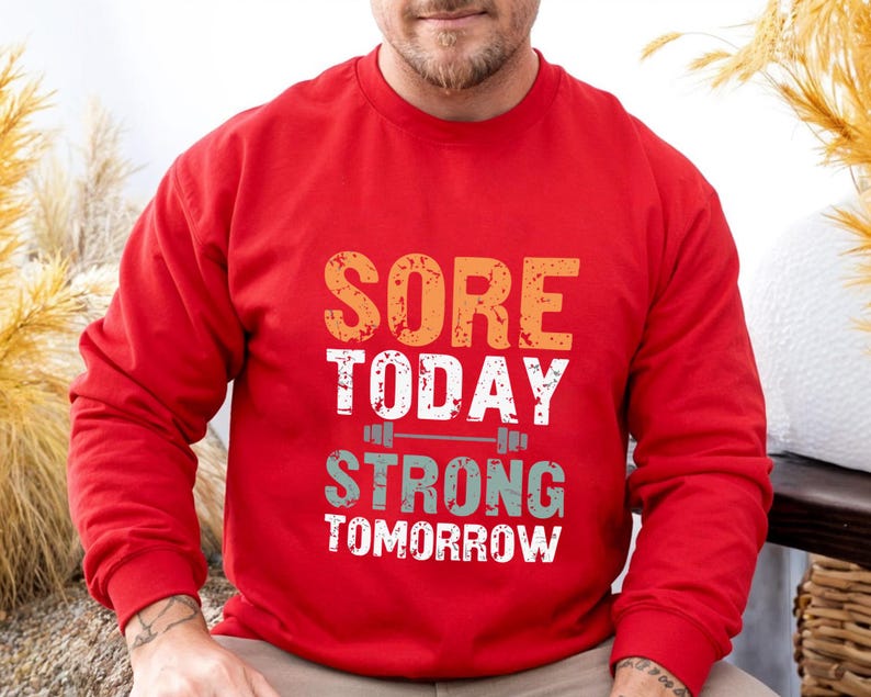 May include: Red long-sleeve sweatshirt with the text "SORE TODAY STRONG TOMORROW" in a vintage style font. The text is layered with a graphic of a barbell.