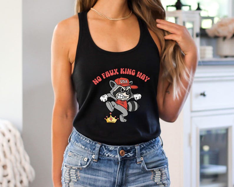 May include: Black tank top featuring a cartoon raccoon wearing a red cap with the text "USA". The raccoon is depicted with an angry expression and is positioned above a gold crown. The text "NO FAUX KING WAY" is arched above the raccoon.