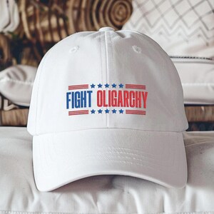 May include: A white baseball cap featuring the text FIGHT OLIGARCHY embroidered in blue and red. The design includes two horizontal red lines above and below the text, with a row of five blue stars centered between the lines.