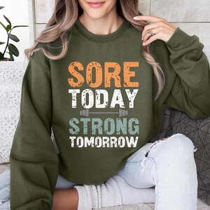 May include: A green sweatshirt with the text "Sore Today Strong Tomorrow" in orange, white, and blue. The text is centered on the sweatshirt and is surrounded by a white outline.