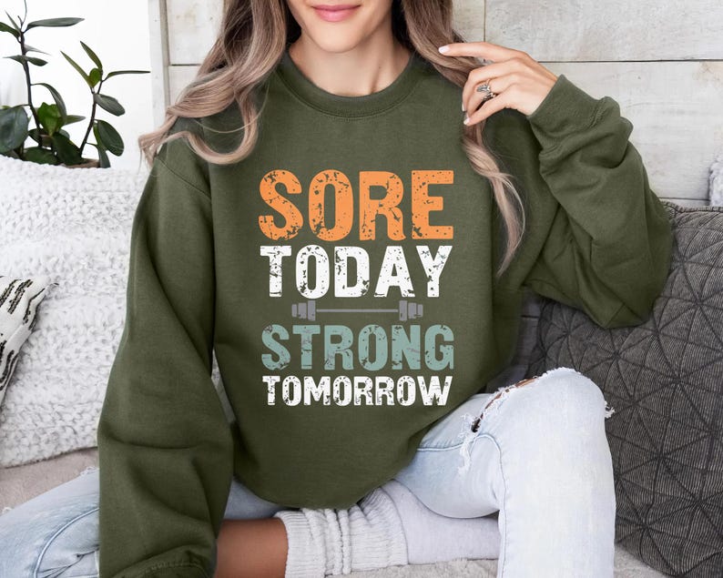 May include: A green sweatshirt with the text "Sore Today Strong Tomorrow" in orange, white, and blue. The text is centered on the sweatshirt and is surrounded by a white outline.