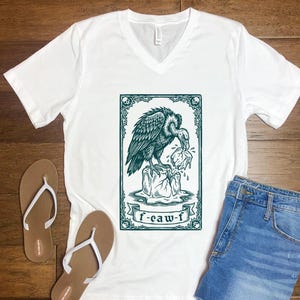May include: A white V-neck t-shirt featuring a dark teal graphic of a vulture perched on a melting ice block. The rectangular frame includes skull accents at the corners and a banner at the bottom with the text f-caw-f in a stylized font.