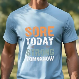 May include: A light blue t-shirt with the text "SORE TODAY STRONG TOMORROW" in orange, white, and green. The text is stylized to look like a distressed, vintage design.