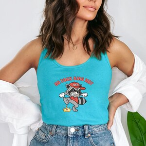 May include: A turquoise tank top featuring a cartoon raccoon wearing a red USA hat, stepping on a gold crown. The text "NO FAUX KING WAY" is arched above the raccoon. The tank top is made of a soft material.